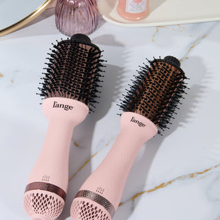 Pink cylindrical hair brushes with black bristles and ergonomic handles, placed on a marble surface.