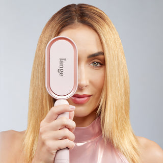 Woman holding a blush L’ange hair tool in front of one eye, showing sleek, straight blonde hair.