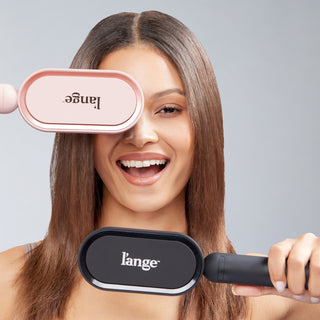 Smiling woman holding two L’ange hair tools, one pink and one black, with sleek straight hair.