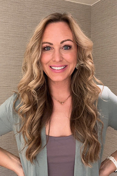 Smiling person with long, wavy blonde hair wearing a light green cardigan and a necklace, standing against a neutral background.