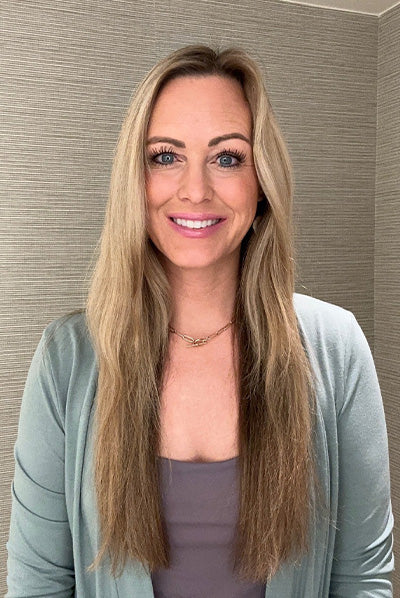 Smiling woman with long blonde hair wearing a light blue blazer and a neutral-colored top, standing against a textured beige background.