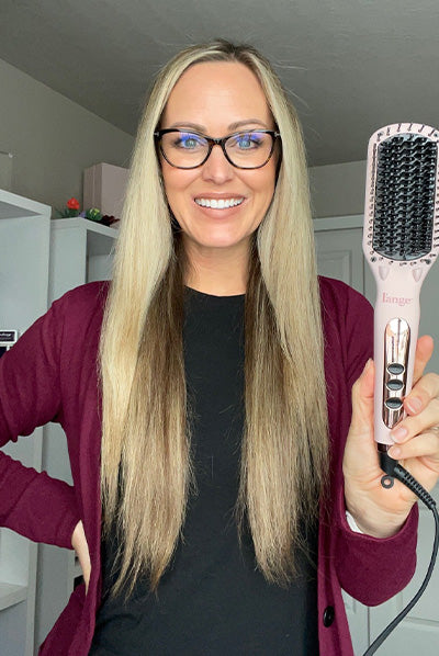 A woman with long, straight blonde hair wearing glasses and a burgundy cardigan holds a pink hair styling tool, smiling in a well-lit room.