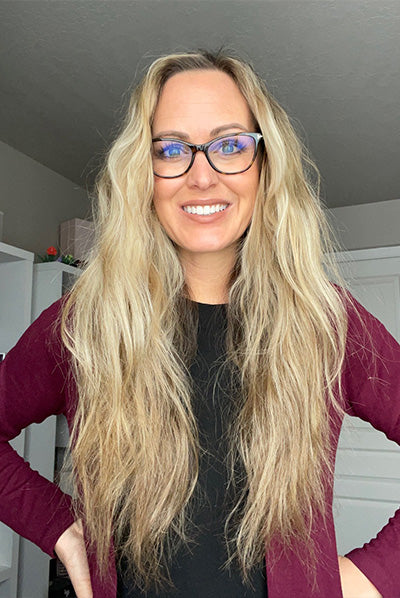 A woman with long blonde hair and glasses smiles while wearing a burgundy cardigan and a black top, standing in a room with white furniture.