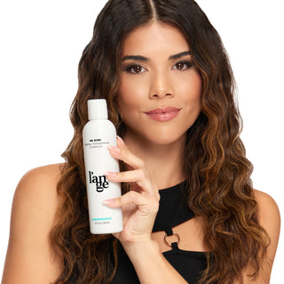 Woman with wavy brown hair holding a bottle of L'ange Ms. Bond Building+ Strengthening Conditioner, displaying sleek styling and product use.