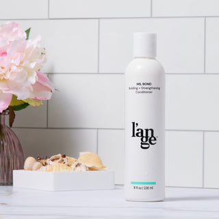 White bottle of l’ange Ms. Bond Building + Strengthening Conditioner with a simple, elegant design, positioned in a stylish bathroom setting featuring decorative pink and white flowers and seashells.