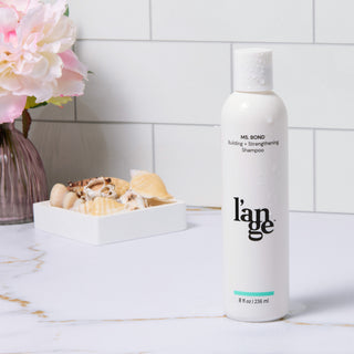 l'ange MS. BOND Building + Strengthening Shampoo displayed on a marble surface, accompanied by seashells and a floral arrangement in the background.