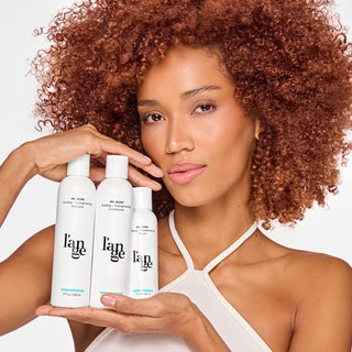 Woman with curly hair showcasing a trio of l’ange hair care products, including building and strengthening shampoo, conditioner, and serum, in sleek white bottles.