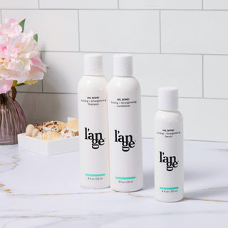 MS. BOND hair care collection featuring building and strengthening shampoo, conditioner, and serum in sleek white bottles. Positioned on a marble surface with a floral arrangement and seashell decor in the background.