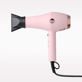A pink hair dryer with a black attachment, featuring a circular control button and a black power cord, shown against a plain white background.