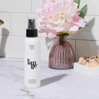 Bottle of L’ange Replenish Detangling Conditioning Spray on a bathroom counter with a pink vase and flower in the background.