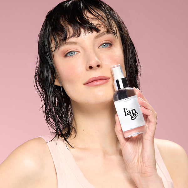 Woman with wet hair holding a bottle of L’ange Rival heat shield spray against a pink background.