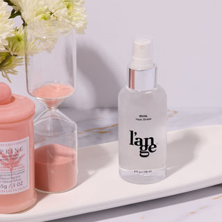 L'ange Rival Heat Shield spray on a marble tray, accompanied by a pink hourglass, floral arrangement, and decorative pink candle.