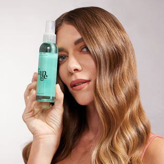 A woman with glossy, honey-blonde waves holds a bottle of Salt + Séa Texturizing Spray next to her face. Her expression is confident, and her hair appears touchably soft and styled in uniform waves.