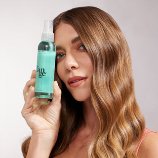 A woman with glossy, honey-blonde waves holds a bottle of Salt + Séa Texturizing Spray next to her face. Her expression is confident, and her hair appears touchably soft and styled in uniform waves.