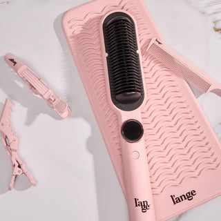 Flat lay of a blush heated paddle brush, silicone mat, comb, and sectioning clips on a marble surface.