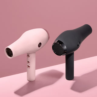 Two L’ange Soleil hair dryers in blush pink and matte black standing upright on a pink surface, showcasing front and back design details including airflow vents and control buttons.