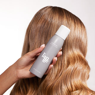 Hand holding a bottle of Style Revival and there's a models side profile of hair that's shiny and has loose waves.