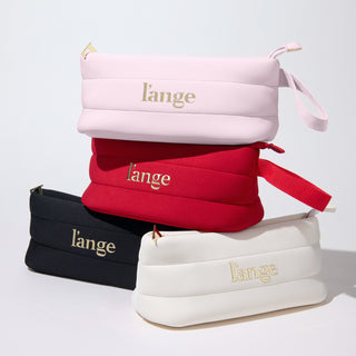 Stacked L’ange zippered cosmetic bags in blush, red, black, and white with gold logos and wrist straps on a white background