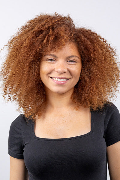 A person with curly, reddish-brown hair smiles while wearing a black square-neck top against a plain background.