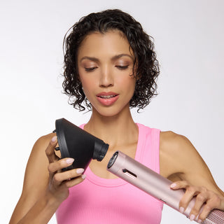 A woman in a pink tank top attaching a black angled diffuser to a rose-gold hair dryer (TurboStyle)