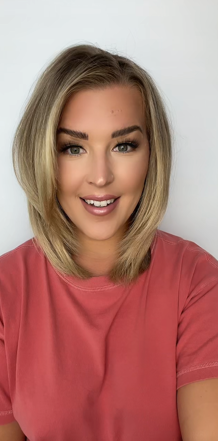 Person with shoulder-length blonde hair wearing a pink shirt, smiling at the camera against a plain background.
