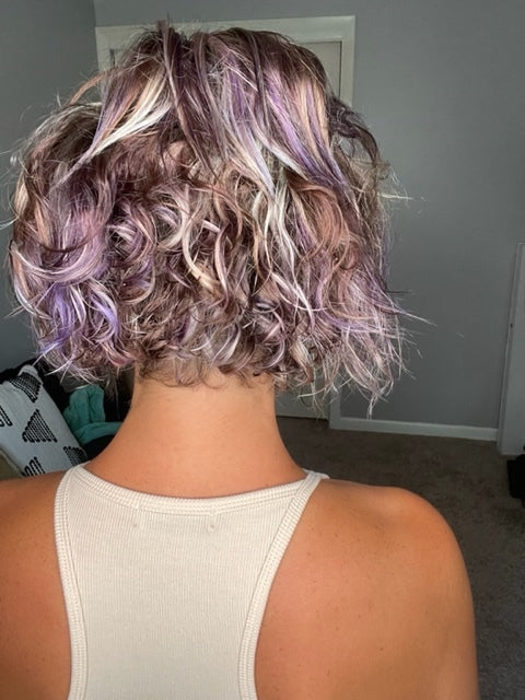 A person with short, curly hair dyed in shades of purple and blonde, wearing a sleeveless beige top, stands facing away in a room with a closed door and carpeted floor.