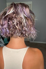 A person with short, curly hair dyed in shades of purple and blonde, wearing a sleeveless beige top, stands facing away in a room with a closed door and carpeted floor.