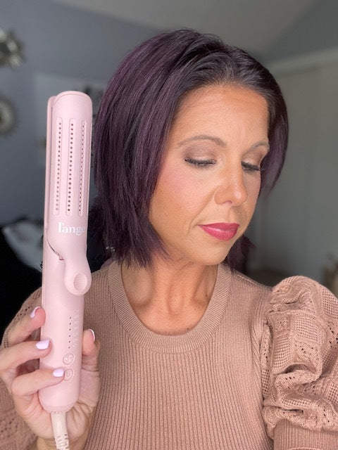 A woman with short dark hair holds a pink hair styling tool, wearing a brown textured sweater and neutral makeup.