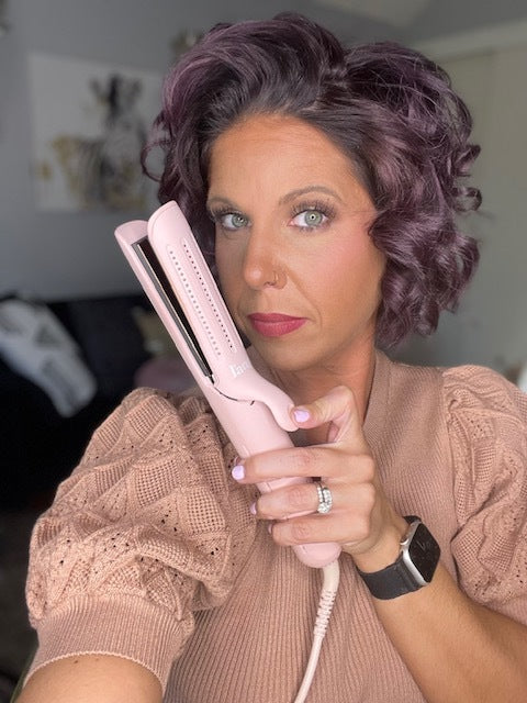 A woman with curled hair holds a pink hair straightener while wearing a beige sweater, posing in a room with a blurred background.