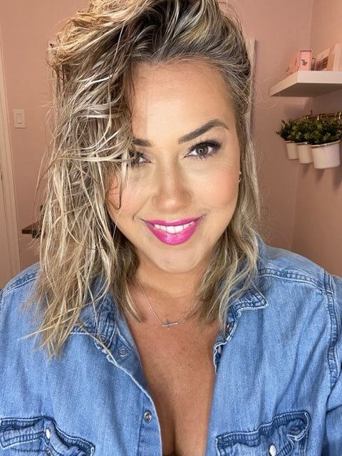 Smiling woman with shoulder-length wavy blonde hair and bright pink lipstick, wearing a denim shirt.