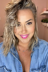 Smiling woman with shoulder-length wavy blonde hair and bright pink lipstick, wearing a denim shirt.
