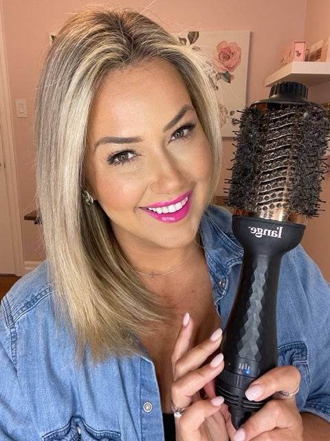 A woman with blonde hair and bright pink lipstick is smiling while holding a large round hairbrush, wearing a denim shirt in a room with pink walls.