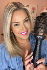 A woman with blonde hair and bright pink lipstick is smiling while holding a large round hairbrush, wearing a denim shirt in a room with pink walls.