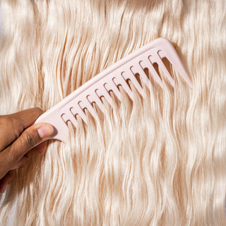A hand gently pulls the pink detangling comb through wavy, platinum blonde hair.